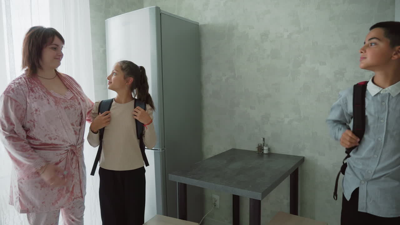 kids in casual wear preparing for school in bright kitchen, mother assists daughter with backpack while brother straps on his bag, notebooks,and water bottles on table, cheerful morning routine