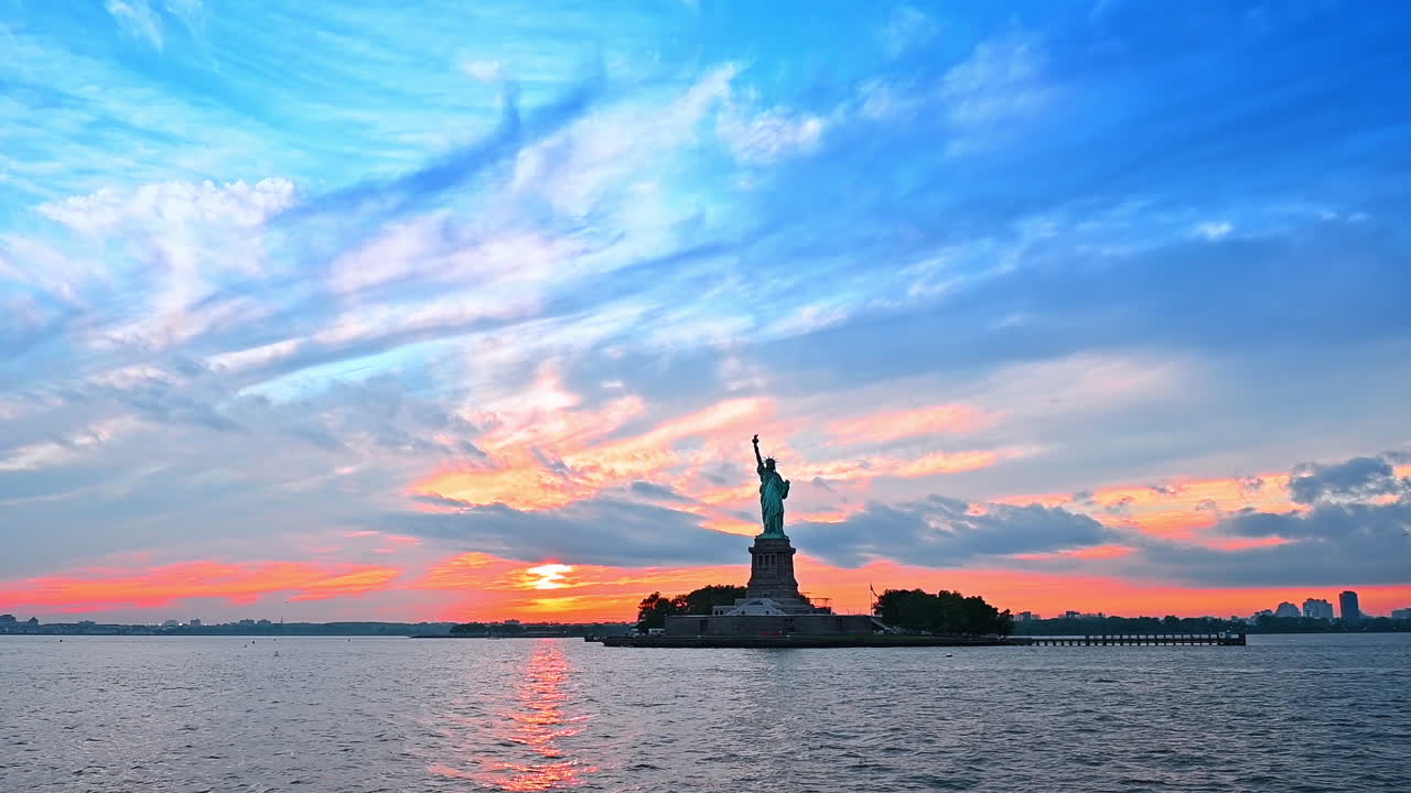 Patung Liberty di Pelabuhan New York. Langit berawan berwarna oranye saat matahari terbenam sebagai latar belakang. Pemandangan dari perairan