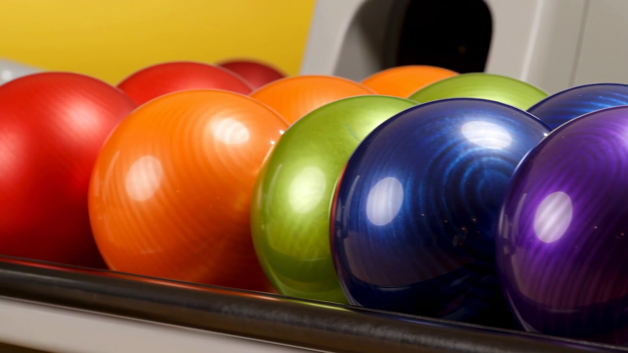 Detailní pohled na těsně seskupené bowlingové koule v zářivě červené, oranžové, zelené, modré a fialové barvě, s leštěnými povrchy, silnými odlesky, malou hloubkou ostrosti a rekreačním vnitřním prostředím