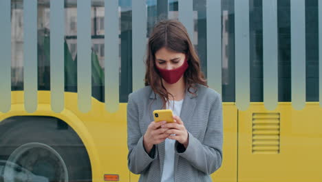 young woman wearing a face mask using smartphone at a bus stop