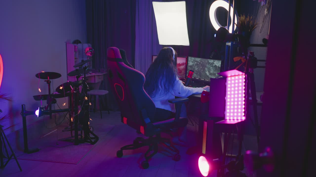 rear view of woman with long hair sitting in gaming chair intensely playing video game on desktop monitors with led lighting gear microphone and headset in dark purple lit room setup