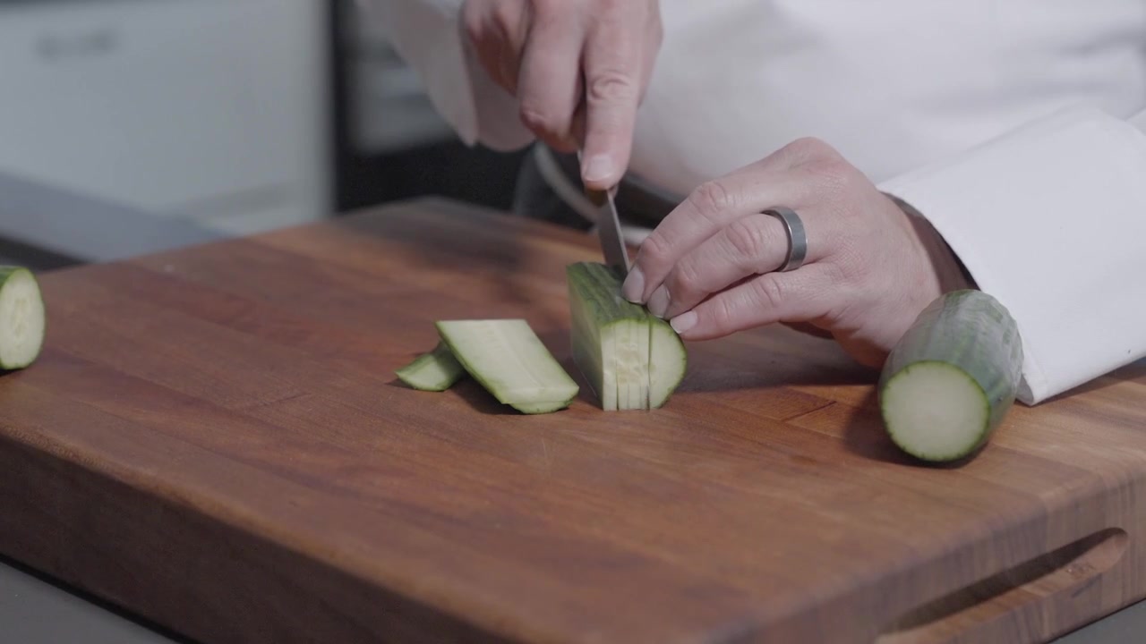 A static shot shows a chef slicing a cucumber lengthwise into thin pieces. Perfect for cooking tutorials, food prep videos, or demonstrating knife skills in culinary content.