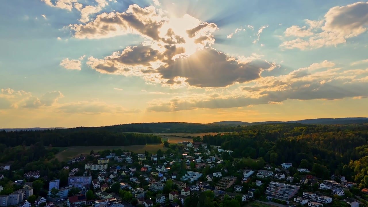 Aerial sunset view over Schaffhausen, Switzerland, with residential neighborhoods, wooded hills and dramatic sunbeams breaking through clouds in warm golden evening light.