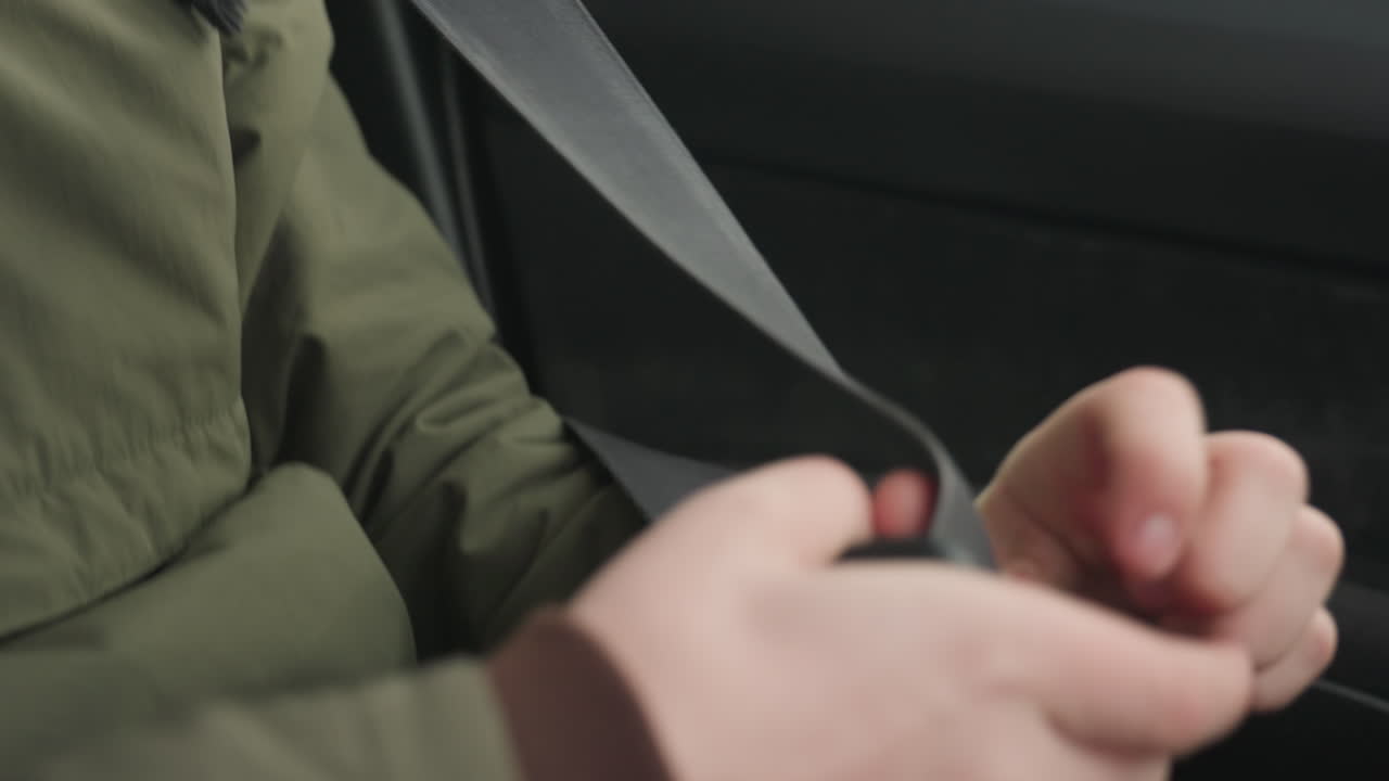 close up of young man in green winter jacket and glasses securing seatbelt near shoulder hook inside car while looking outside window with blurred city buildings visible in cold snowy background