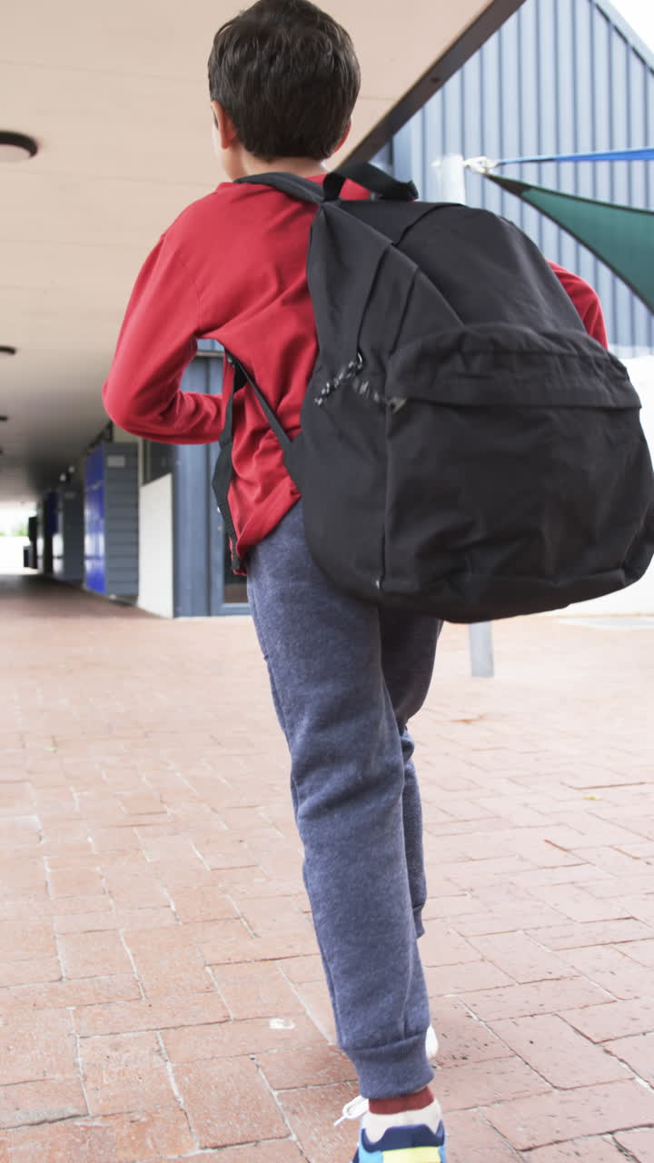Vertical video: In school setting, young biracial student walks with black backpack