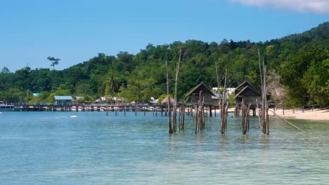 Panorámica-Lenta-De-Una-Pintoresca-Isla-Tropical-Exótica-Con-Playa-De-Arena-Blanca,-Cabañas-De-Madera-En-La-Playa-Con-Vistas-Al-Agua-Turquesa-Del-Océano-En-Una-Isla-Cubierta-De-árboles-De-Selva-Tropical-En-Raja-Ampat,-Papua-Occidental,-Indonesia