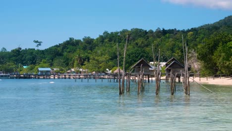 una lenta panela de escénica isla tropical exótica con playa de arena blanca, cabañas de playa de madera con vistas al agua turquesa del océano en la selva tropical cubierta de árboles isla en raja ampat, papúa occidental, indonesia