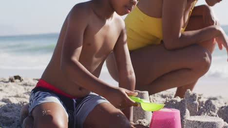 smiling african american son with mother building sandcastle on sunny beach