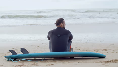 back view of a sportsman with artificial leg sitting on sandy beach and resting after surfing workout in ocean