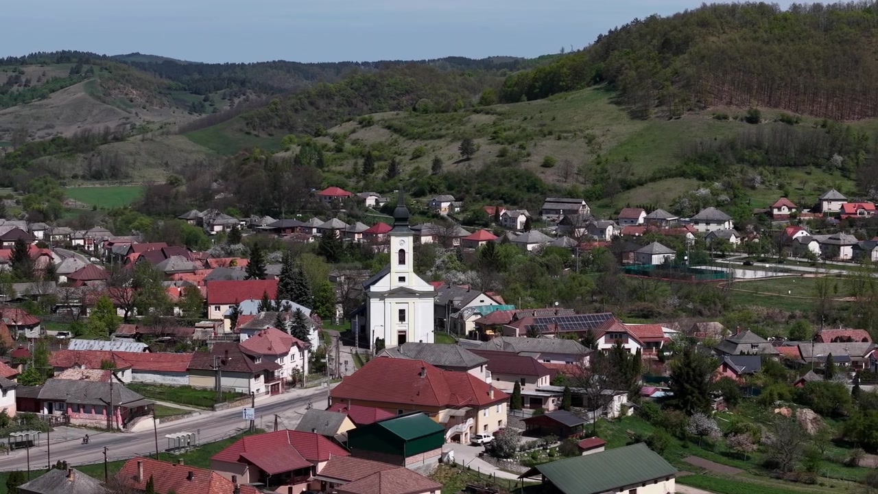 Zvýšená perspektiva odhaluje malebnou maďarskou vesnici, její tradiční domy, centrální kostel a okolní zelené kopce, ztělesňující klidnou venkovskou atmosféru.