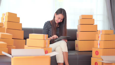 work from home concept - young asian lady making a delivery list using a tablet computer for shipping parcel boxes from her home, working at home