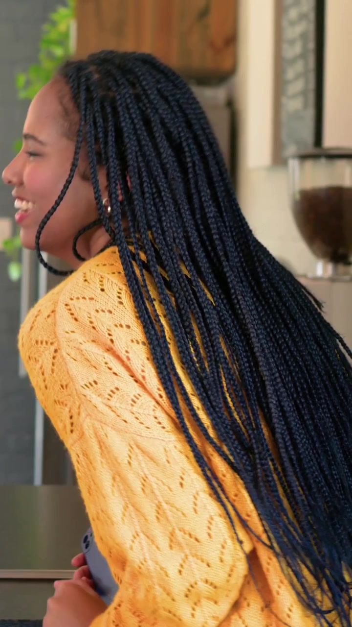 Donna che parla con un maglione di maglia giallo, gesticola e si sistema la treccia al bancone di un caffè con uno smartphone
