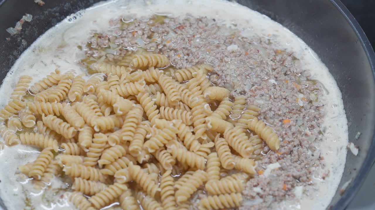 Close-up hand pouring dry fusilli pasta into bubbling creamy meat sauce with minced beef in pan