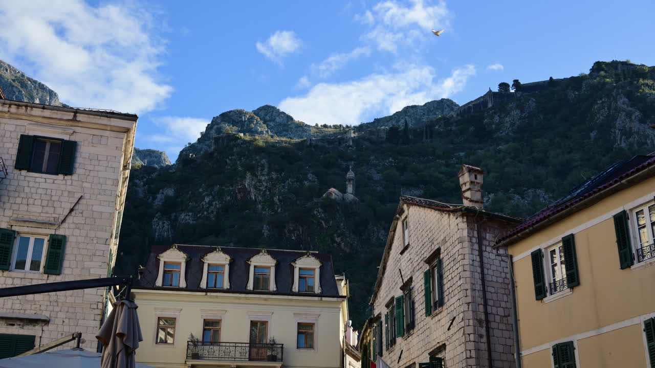 Edifici in pietra con persiane che si stagliano di fronte ad aspre montagne costiere sotto un cielo azzurro.