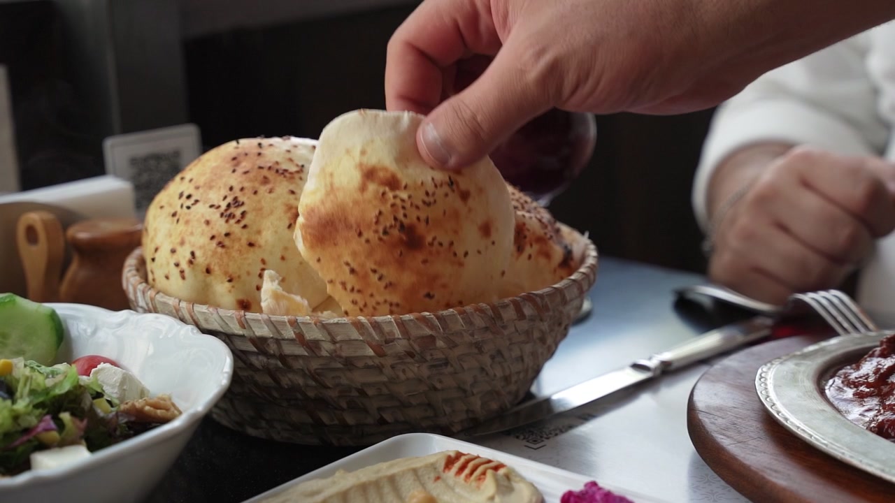 Close up of a man's hand picking up a piece of puffed pita bread from a wicker basket. Served with hummus and salad at a Turkish restaurant during lunch.