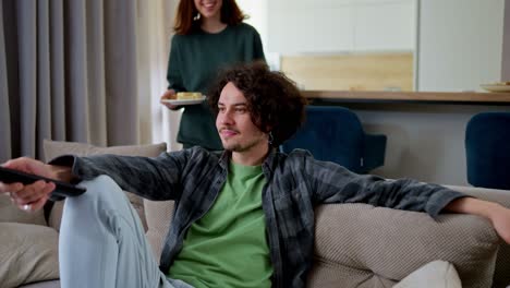 Happy-brunette-guy-with-curly-hair-in-a-gray-checkered-shirt-watches-TV-while-he-is-brought-sweet-snacks-by-his-brunette-girlfriend-in-a-modern-apartment-at-home