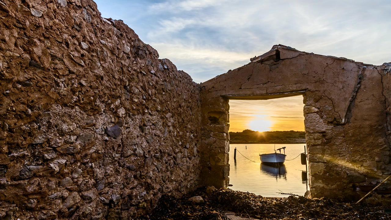 Pequeno barco flutuando na entrada das ruínas romanas do porto de Sanitja sob o pôr do sol dourado de Menorca em timelapse.
