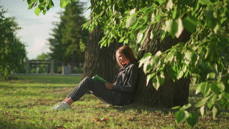 dama sentada al aire libre bajo un árbol en un campo de hierba, leyendo un libro con la cálida luz del sol filtrando a través de las hojas, creando un ambiente pacífico, rodeada de vegetación y naturaleza