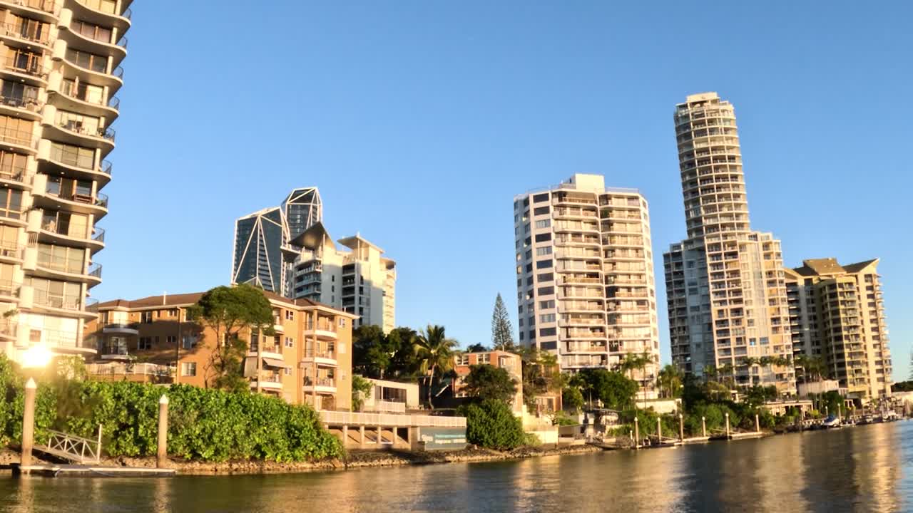 A serene view of modern high-rise buildings along a tranquil riverbank under a clear blue sky.
