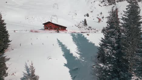 zipline drone shot of a log cabin beside a frozen pond and right next to a snowy mountain slope, located at engelberg, brunni, in bahnen, switzerland