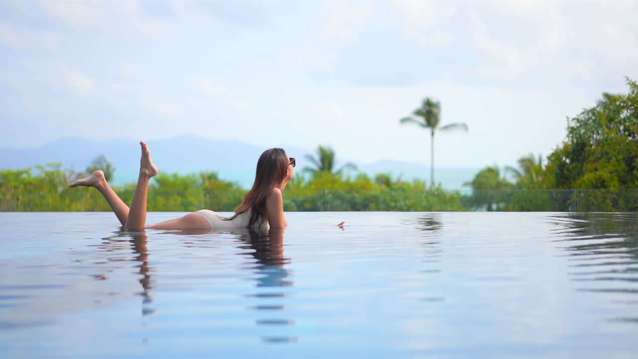 Attraktive asiatische Frau liegt im flachen Wasser eines Infinity-Pools mit atemberaubender Aussicht auf tropische Vegetation, statische Zeitlupe.