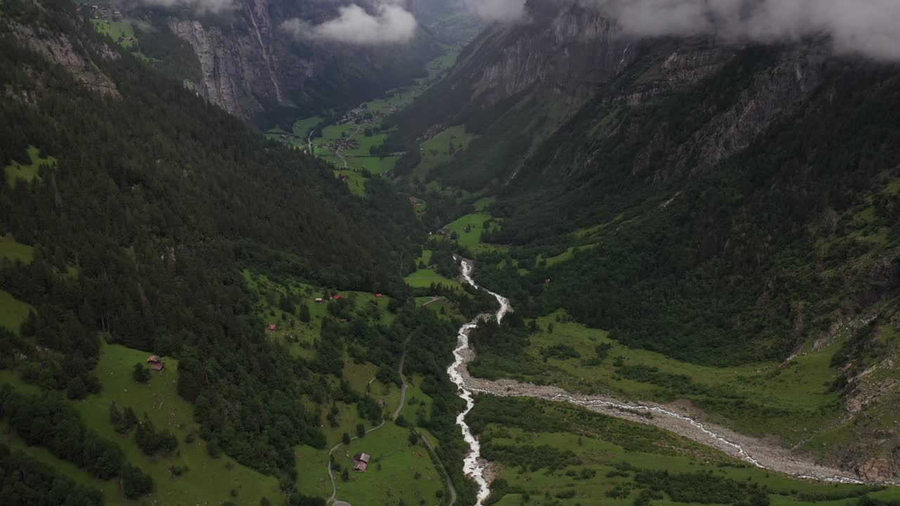 An drone shot moving forwards and tilting up, revealing a small creek running down from the mountain to the small village, surrounded by beautiful and lush greenery in the summer in Lauterbrunnen