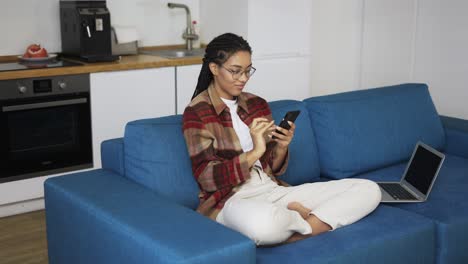 Junge-Frau-Benutzt-Smartphone-Zu-Hause-Auf-Einer-Couch-Mit-Laptop