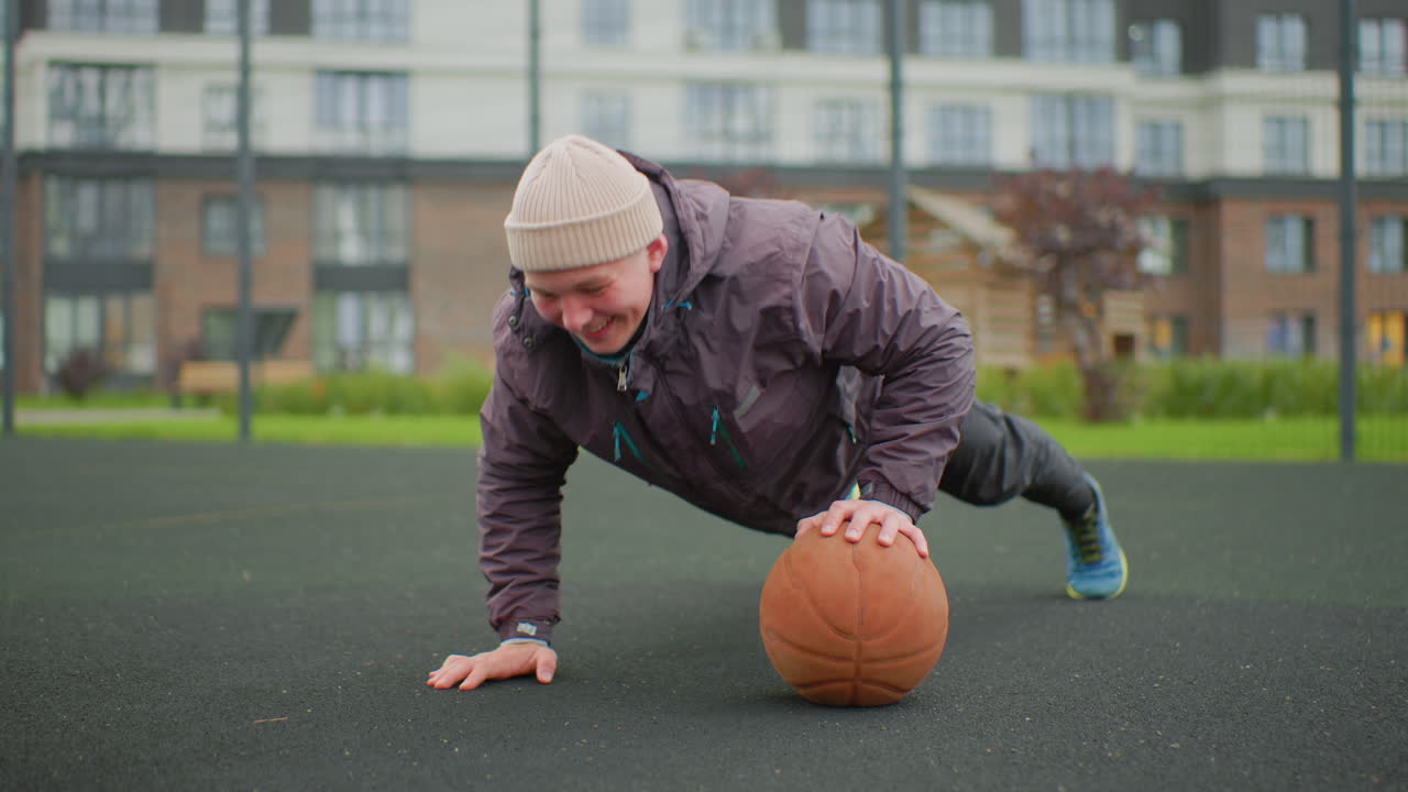 Uomo anziano che cerca di raggiungere un pallone da basket all'aperto su un campo da quartiere in gomma, che indossa un berretto e un piumino mentre l'allenatore lo aiuta a mantenere l'equilibrio durante un push-up in stile riabilitazione, dimostrando resilienza