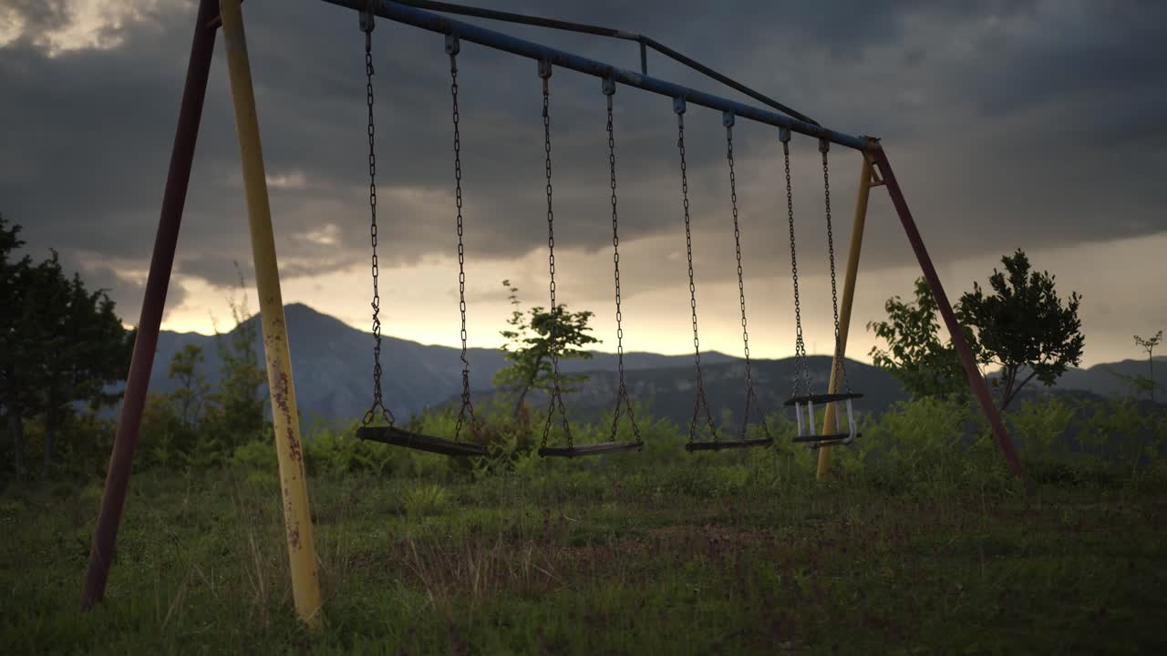 Static wide shot of an old metal swingset with multiple empty seats, standing in an overgrown field with distant mountains and dramatic evening clouds.
