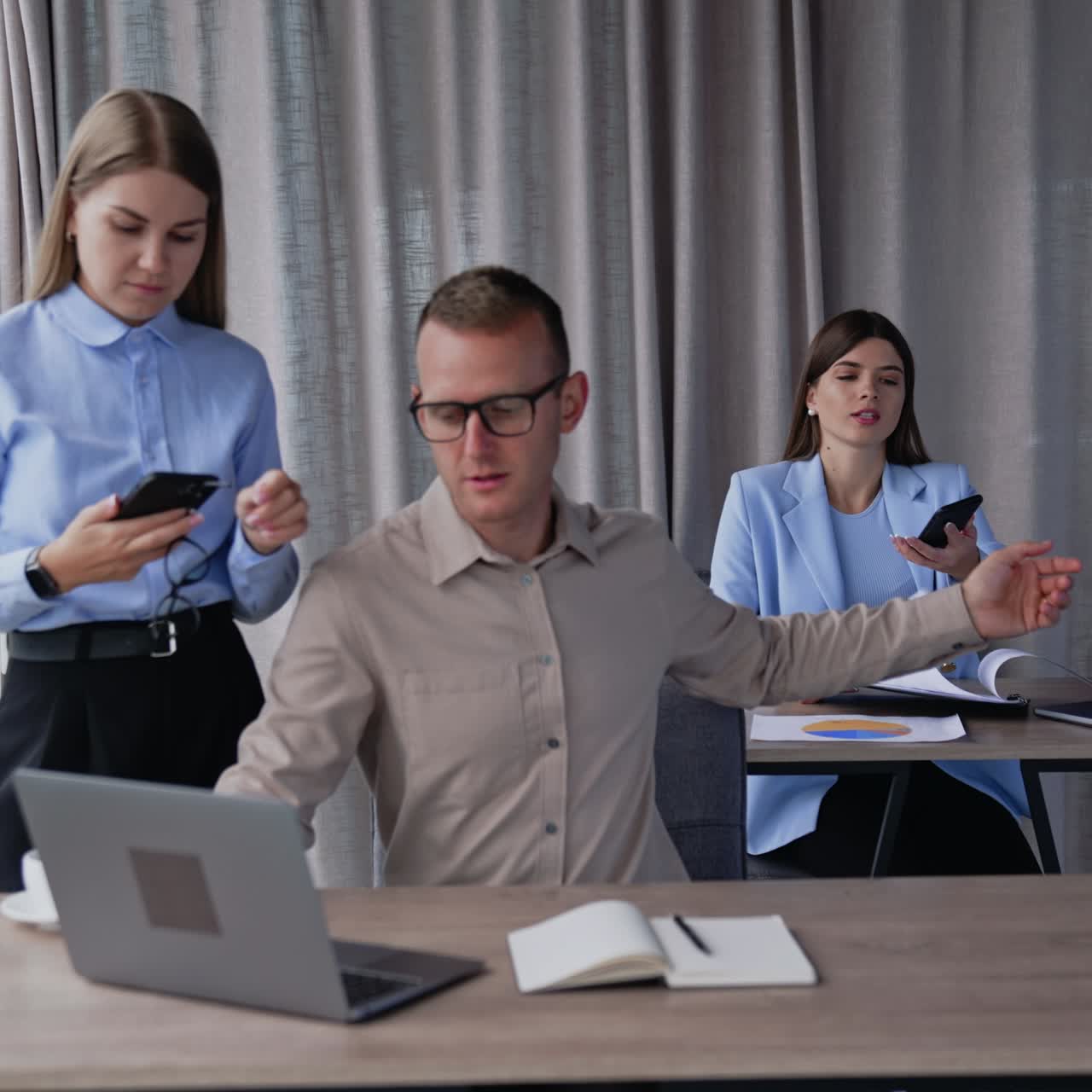Team of colleagues in the light office room. Man sitting at computer takes phone and passes it to female teammate. Blonde lady with phone in hands standing beside male employee
