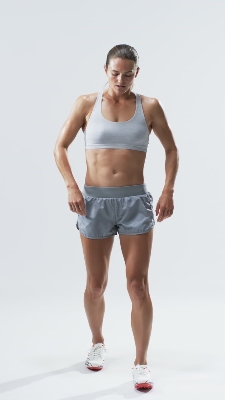 Vertical video: Caucasian female athlete standing, looking down, white background