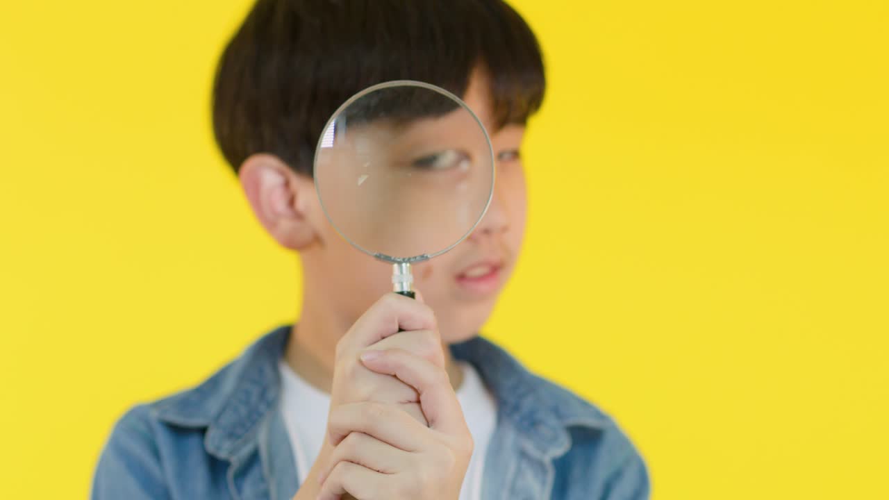 Smiling boy in denim jacket explores with magnifying glass, bright yellow backdrop, playful close-up