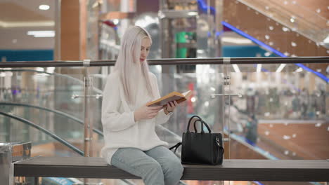 youthful woman reading novel on mall bench by glass elevator, wearing casual jeans and white sweater while carrying stylish black purse, serene calm moment of focused concentration and leisure