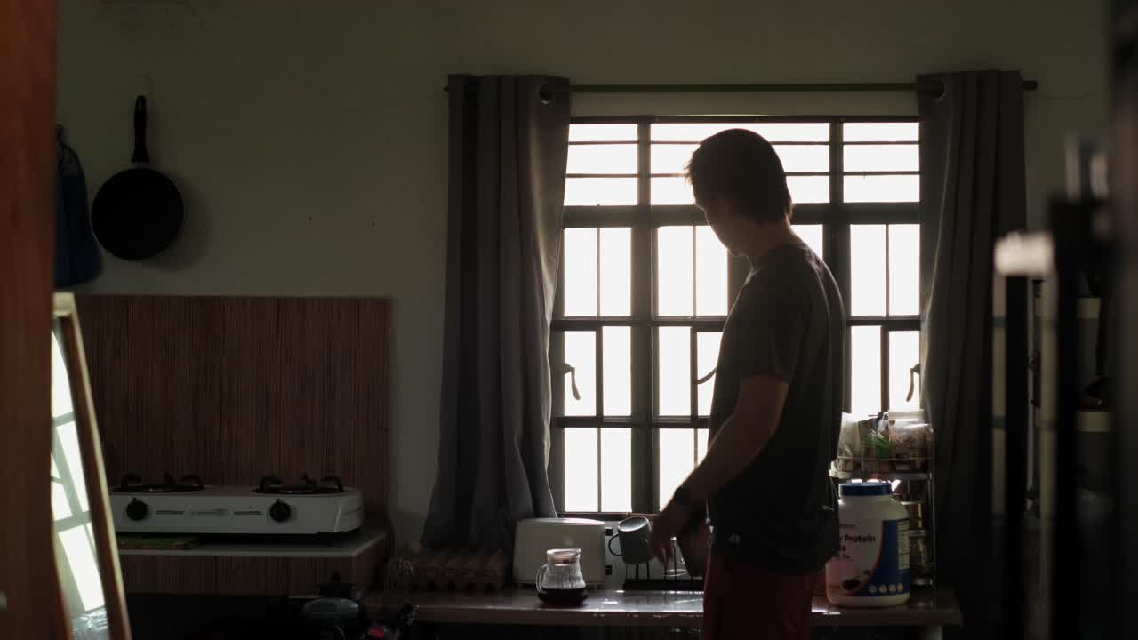 Young man shaking protein shake bottle and drinking by sunlit apartment window - wide static shot