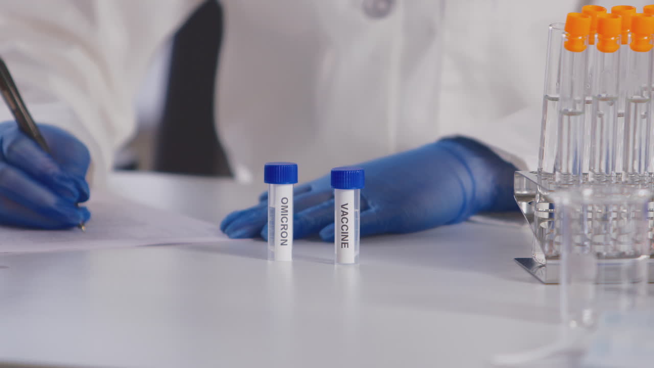 Close Up Of Female Lab Research Worker In PPE Studying Test Tubes Labelled Omicron And Vaccine