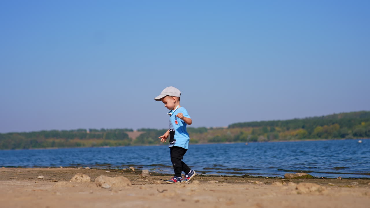 Baby boy walks by the river waterfront on sunny day. Caucasian toddler looking for stones at the beach.