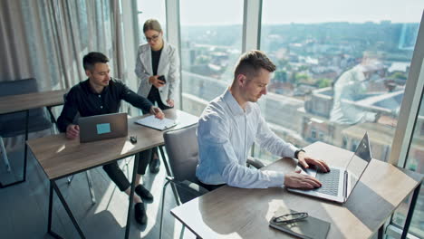 office employees working in a sunny modern office. men sitting at desk in front of laptops. female in white jacket standing near the male colleague in black shirt.