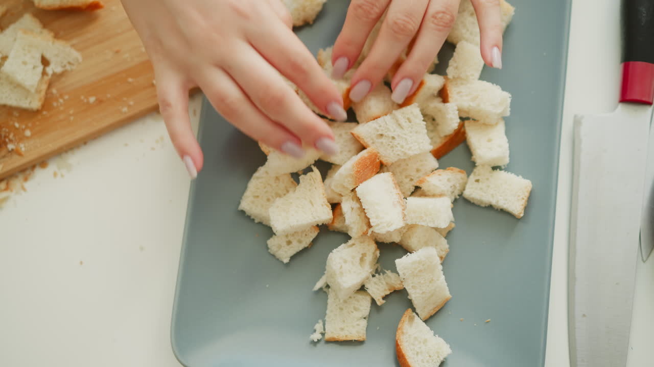 Saucier hand pouring chopped bread cubes into plastic pan on white table next to knife and cutting board, crumbs scattered under soft window light, capturing precise calm culinary prep moment