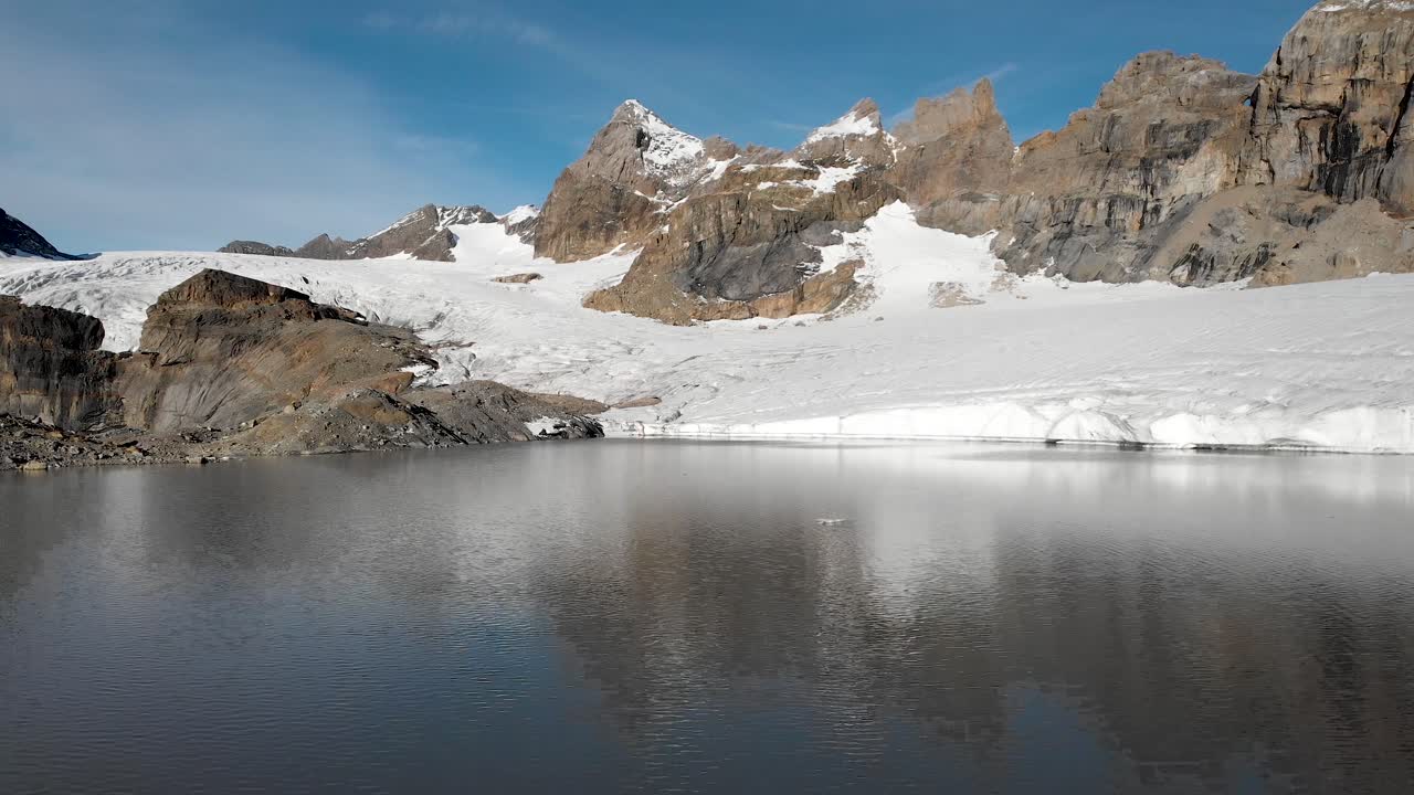 Aerial flyover over the water and icebergs of glacial lake towards Claridenfirn glacier in Uri, Swizerland on a calm, peaceful morning with alpine peaks behind a wall of ice
