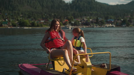 woman and kid in life jackets rest actively in catamaran. young mom smiles moving pedals and daughter looks back at shore with interest slow motion