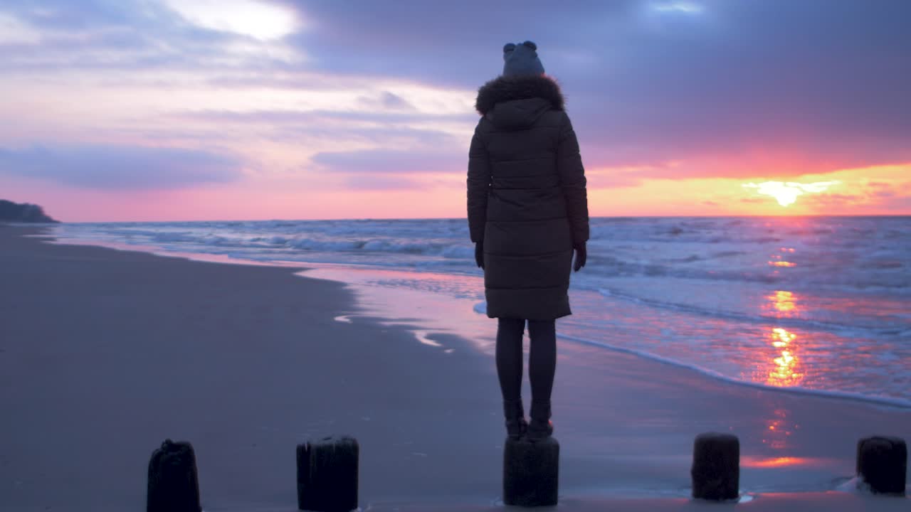 겨울 옷을 입은 젊은 여성이 오래된 목재 부두, 발트해 해변의 모래사장에 서서 낭만적인 일몰을 바라보며 중간 추적 샷 오른쪽