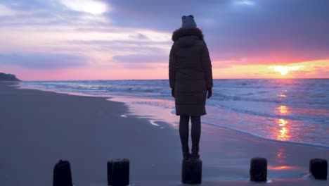 young woman in winter clothes standing on old wooden pier, sandy shore of the baltic sea beach, watching romantic sunset, medium tracking shot right