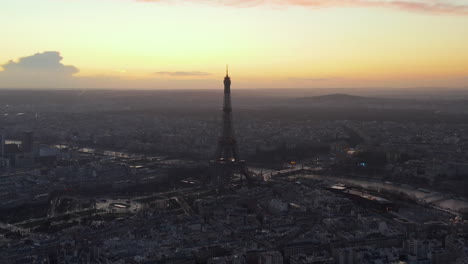 Aerial-descending-footage-of-dominant-tourist-landmark-in-large-city-at-dusk.-Eiffel-Tower-and-colour-sunset-sky-in-background.-Paris,-France