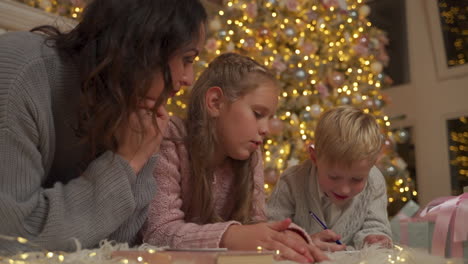 little boy writes a letter with his sister and mother on the floor next to the gifts and the christmas tree