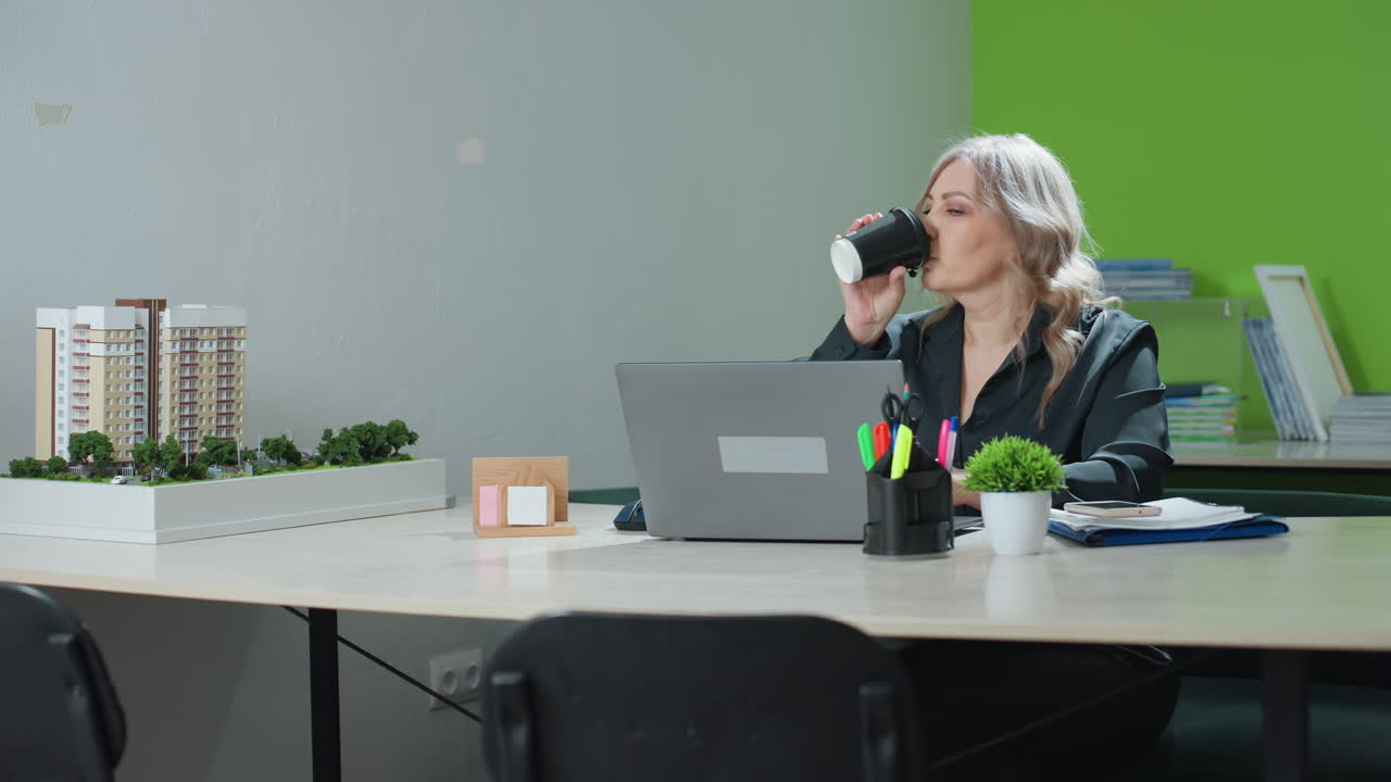 Real estate marketer seated at desk focuses on laptop while working on task, pauses to sip takeaway coffee and continues, surrounded by office decor, building model and folders