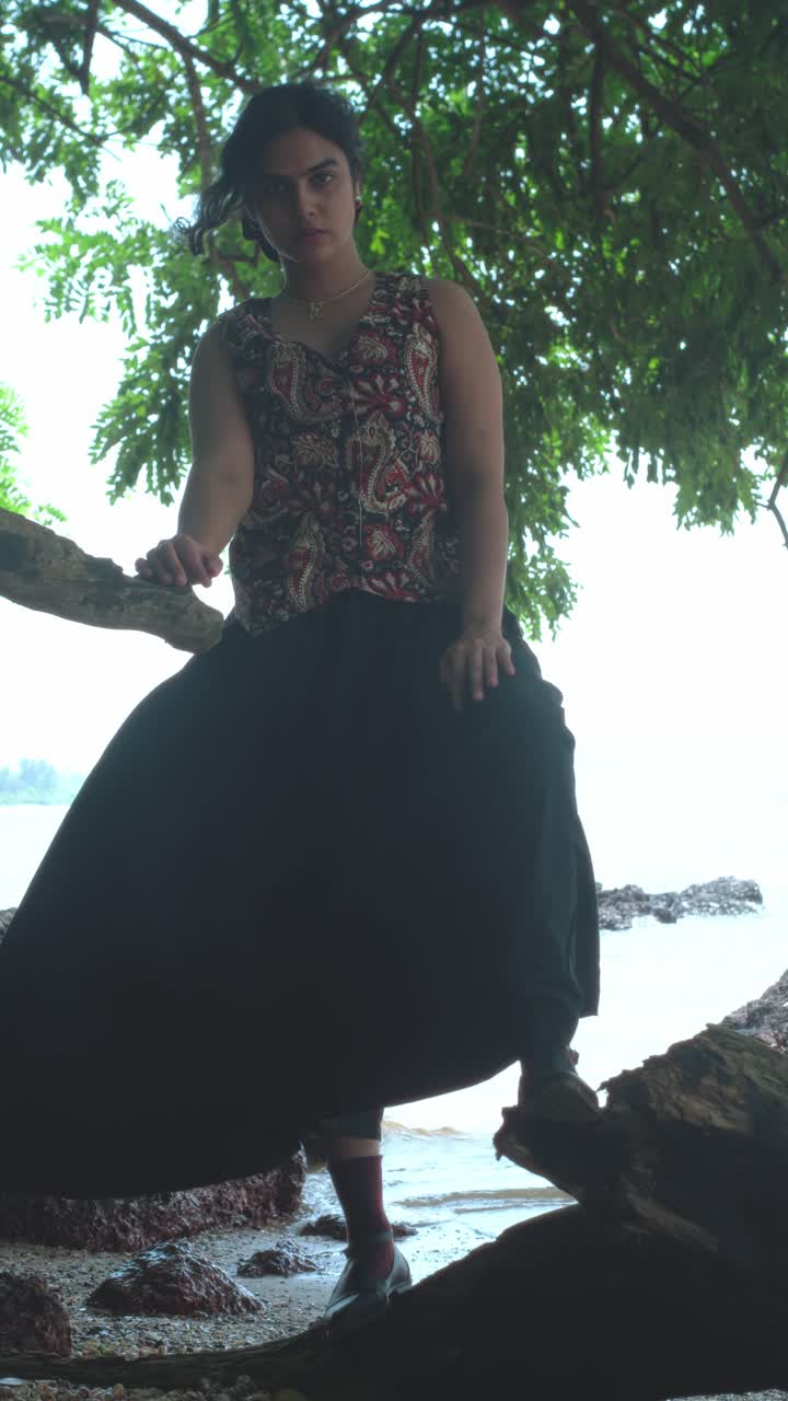 Toma vertical de una mujer subida a la rama de un árbol sobre la costa con olas