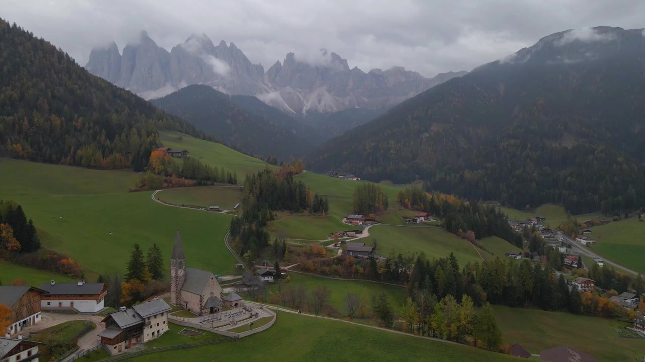 Luftfoto af Santa Maddalena kirken i Val di Funes med farverigt efterårsløv og Odle-bjergene dækket af skyer i baggrunden, Sydtyrol, Italien, drone trækker sig ud