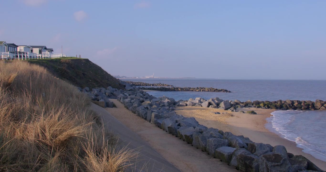 Inquadratura zoom extra ampia di scogliere frangiflutti, diga marina ed erba delle dune, presso il villaggio vacanze di Hopton