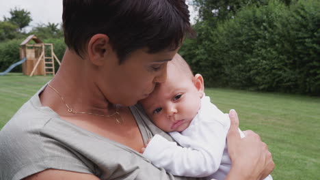 loving mother holding and kissing 3 month old baby daughter in garden at home