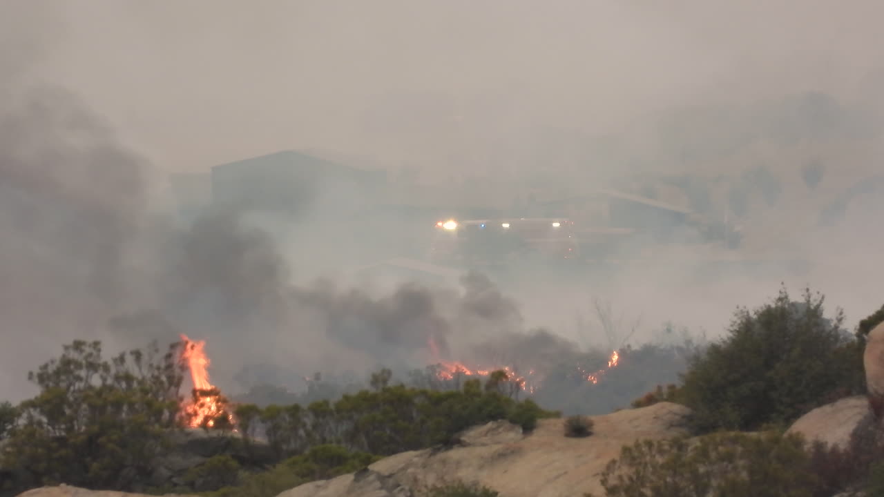 los arbustos en llamas durante el incendio de fairview en hemet california, con las luces intermitentes de los servicios de emergencia visibles a través del humo feroz en el fondo